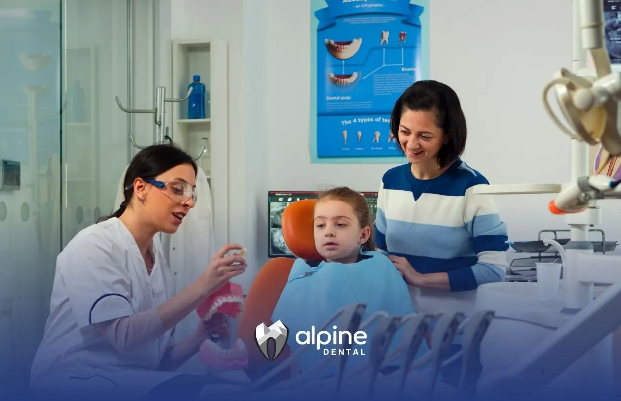 Child receiving dental care while a dentist explains treatment steps to a young girl and parent in a modern clinic setting.